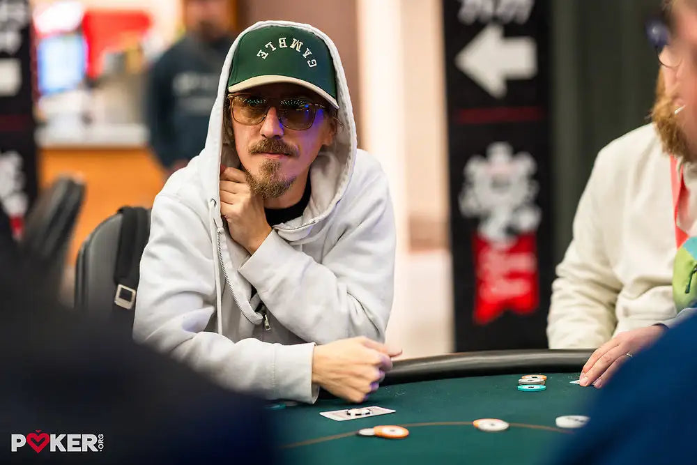 A man wearing a green and white ’say yes’ baseball cap with sunglasses, a light gray hoodie, and a goatee sits at a poker table.